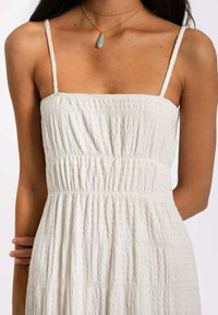Robe blanche texturée avec de fines bretelles et des détails froncés au niveau du corsage. Un collier en or à couches avec un pendentif en turquoise est visible.