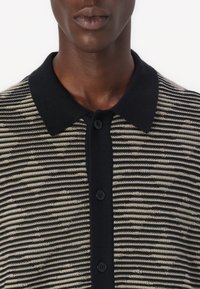 Strikket polo shirt med stripemønster i beige og svart, med svart krage og knappelukking, glatt tekstur og klassisk design.