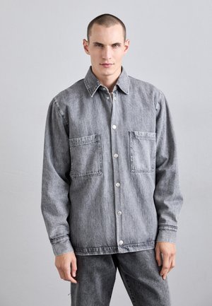 Grå denimjakke med spidse krave, frontknapper og to store lommer. Har en afslappet pasform og detaljering med stikninger.