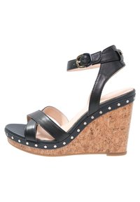 Czarne skórzane sandały na koturnie z paskiem wokół kostki i złotym klamrą, korkowa podeszwa z białymi nitami oraz teksturowana podeszwa.