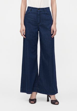 Παντελόνι denim με σκούρο μπλε χρώμα και φαρδιά πόδια, φορεμένο με μαύρα ψηλοτάκουνα πέδιλα και λευκή μπλούζα, εμφανίζεται από τη μέση μέχρι τα πόδια.