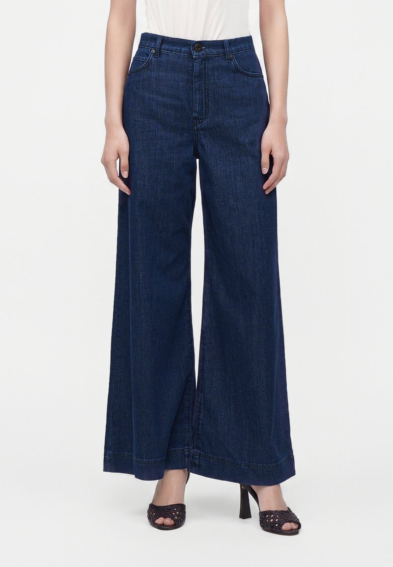 Παντελόνι denim με σκούρο μπλε χρώμα και φαρδιά πόδια, φορεμένο με μαύρα ψηλοτάκουνα πέδιλα και λευκή μπλούζα, εμφανίζεται από τη μέση μέχρι τα πόδια.