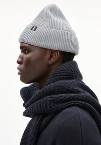 Jongere in profiel met een grijze gebreide beanie en een dikke, donker gebreide sjaal om de nek over een donkere trui.