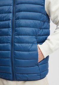 Person trägt eine blaue, gesteppte Weste mit Reißverschluss, mit der Hand in der Seiten-Tasche, über einem cremefarbenen Langarmoberteil, vor einem einfarbigen Hintergrund.