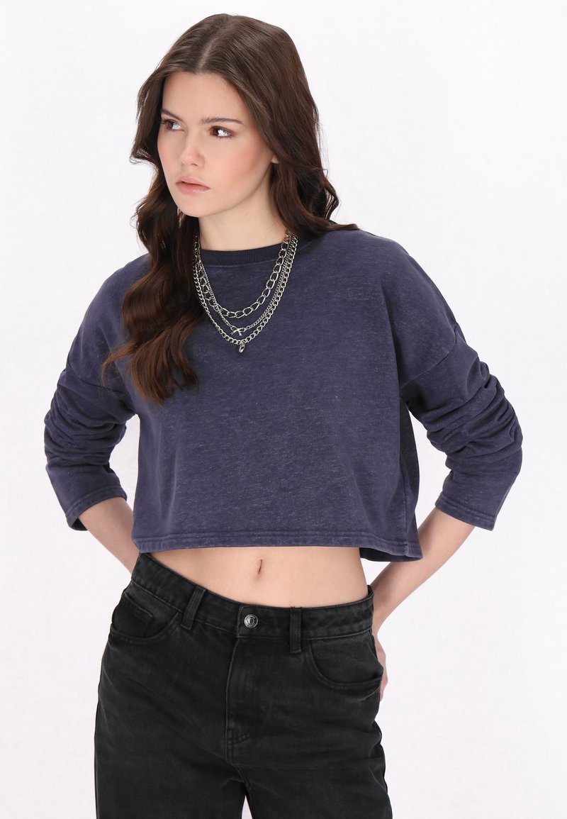 Granatowy krótki sweter z długimi rękawami, o miękkiej fakturze i okrągłym dekolcie. Połączony z czarnymi jeansami o wysokim stanie i warstwą srebrnych łańcuszków.