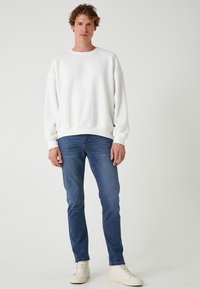 Felpa bianca in cotone con vestibilità comoda, polsini a costine, indossata sopra jeans blu slim fit e sneakers bianche. Design semplice e pulito.