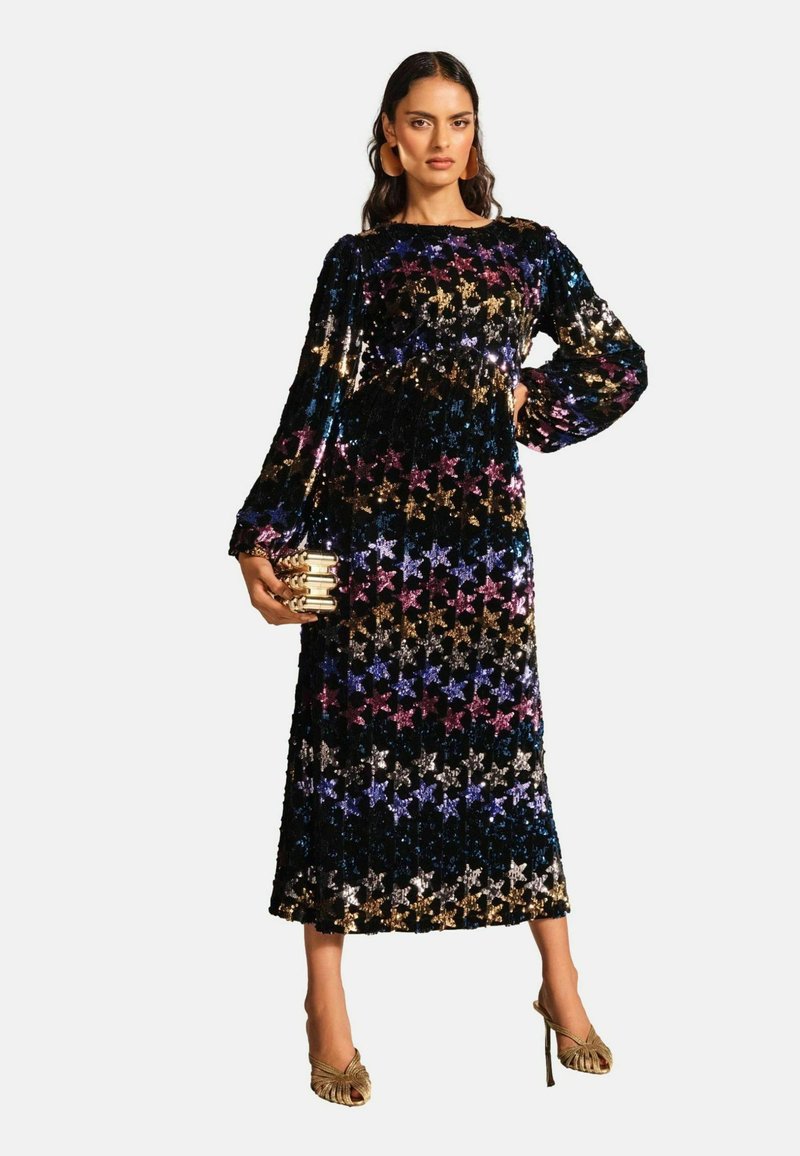 Rochie lungă cu mâneci lungi, decorată cu strasuri, având un model de stele multicolore pe un fundal negru. Modelul poartă o clutch aurie și tocuri cu siret.