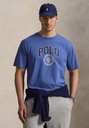 Uomo che indossa una maglietta Polo Ralph Lauren blu e un cappellino, pantaloni della tuta grigi e un maglione blu navy legato intorno alla vita.