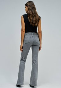 Mujer con cabello largo castaño, vistiendo una blusa negra sin mangas y unos pantalones de mezclilla de tiro alto grises, de pie de espaldas sobre un fondo liso.