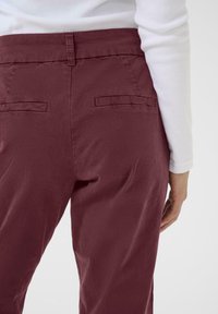 Maroon trousers with a smooth texture, featuring back pockets and a standard waistband. Worn with a long-sleeve white top.