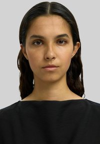 Schwarzes, strukturiertes Kleidungsstück mit weitem Ausschnitt. Das Subjekt hat lange, dunkelbraune Haare und einen neutralen Hintergrund. Neutrale Gesichtsausdruck.