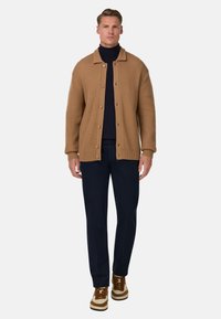 Bruine geribde cardigan met knoopsluiting, gelaagd over een marineblauwe coltrui. Gecombineerd met marineblauwe broek en tan en witte sneakers.