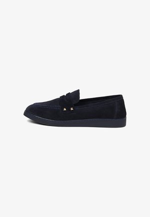 Navyblauwe suède loafer met decoratieve band, contrasterende stiksels en platte rubberen zool, zijaanzicht op witte achtergrond.