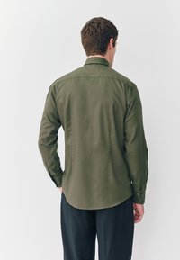 Camisa de manga larga verde oliva con cuello recto y puños con botones, presenta una textura suave y un dobladillo curvo.