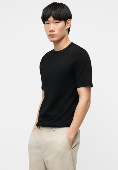 Νεαρός άνδρας με μαύρο κοντομάνικο πουκάμισο και μπεζ παντελόνι, στέκεται με τα χέρια στις τσέπες μπροστά σε απλό φωτεινό φόντο.