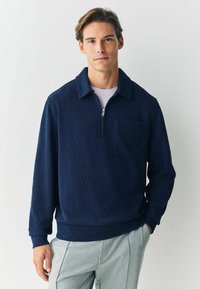 Giovane uomo con un pullover zip-up blu navy e tasca sul petto, indossando pantaloni grigio chiaro, in piedi davanti a uno sfondo bianco semplice.