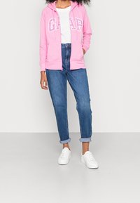 Sudadera rosa con cremallera y logo "GAP", combinada con jeans de lavado medio y puños enrollados, y zapatillas blancas. El modelo se presenta frente a un fondo neutro.