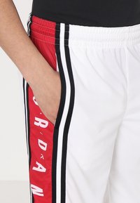 Pantalons de sport blancs avec des panneaux latéraux rouges portant le texte "JORDAN". Des rayures noires mettent en valeur le design. Comprend des poches et une ceinture élastique.