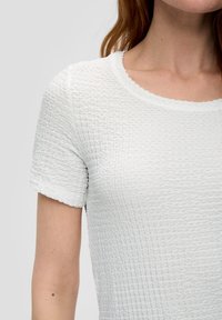 Haut blanc texturé avec des manches courtes et un col festonné, présentant un motif froncé sur tout le tissu. Fini lisse au niveau du col.