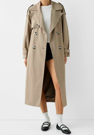 Frau trägt einen beigen, doppelt geknöpften langen Trenchcoat über einem weißen Top, schwarzen Shorts, weißen Socken und schwarzen Mary Jane Schuhen.