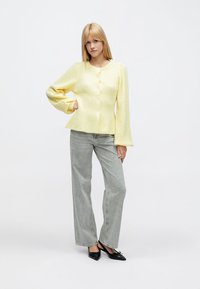 Geltonas ribuotas megztinis su ilgomis, plačiomis rankovėmis ir priekinėmis sagomis, derinamas su šviesiai pilkais plačiomis kelnėmis ir juodais lygiais batais.