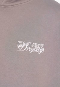 Svetlo hnedá látka s bielym prekryvným textom, ktorý znie "DRPSZ Dropsz" v tučných a kurzívnych písmach.