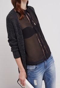 Grå strikket cardigan over en gennemsigtig sort skjorte med knaplukning foran, sorte blonder detaljer, parret med slidte blå jeans og en hvid clutch.