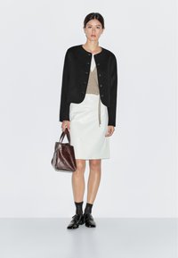 Kvinna stående i svart knäppt jacka, beige topp, vit läderkjol, svarta strumpor, svarta skor, håller en brun läderhandväska.