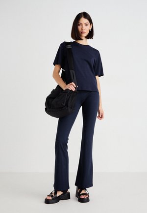 T-shirt marine, pantalon évasé et sandales noires. Le modèle tient un sac noir avec des accents en maille. Design simple, texture lisse, peu de détails.