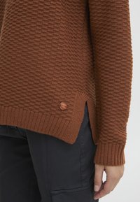 Pull en tricot marron avec un motif texturé, des poignets côtelés, des fentes sur les côtés et un petit accent en bois près de l’ourlet.