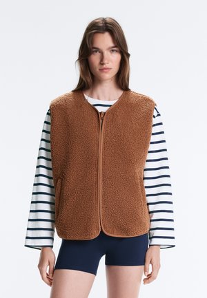 Jeune femme portant un gilet en polaire brun zippé par-dessus une chemise blanche à manches longues avec des rayures noires et un short bleu marine, debout devant un fond uni.