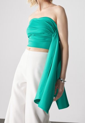 Femme portant un top turquoise sans bretelles avec un tissu drapé et un pantalon blanc à jambes larges, accessoirisée d’un bracelet en argent au poignet gauche.