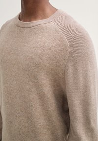 Beige stickad tröja med rund halsringning och raglanärmar, med en mjuk textur och subtila färgavvikelser.