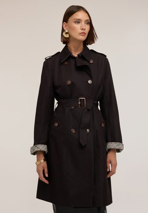 Frau mit kurzen braunen Haaren trägt einen schwarzen Trenchcoat mit Gürtel und hochgekrempelten karierten Manschetten, große goldene Creolen und goldene Armbänder an einem Handgelenk.