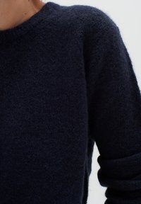 Lähivõte inimesest, kes kannab tume tumesinist karvase pehme materjaliga kampsunit, millel on ümmargune kaelus ja pikad varrukad, tasase heleda tausta ees.