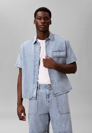 Mann, der ein hellblaues kurzärmeliges Jeanshemd über einem weißen T-Shirt mit passenden hellblauen Jeans-Shorts trägt, vor grauem Hintergrund stehend.