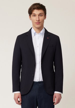 Blazer marine en tissu lisse avec deux boutons, poches latérales et un petit logo rouge. Porté par-dessus une chemise blanche à boutons.