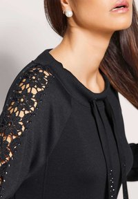 Frau trägt ein schwarzes Oberteil mit floralen Spitzen-Ausschnitten an den Ärmeln und Perlen-Ohrsteckern, mit langen braunen Haaren.