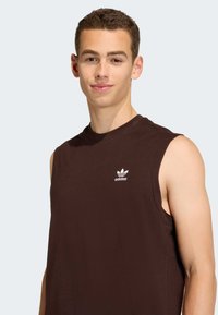 Hnědé tílko z bavlny s bílým logem Adidas na hrudi, hladkou texturou a kulatým výstřihem.