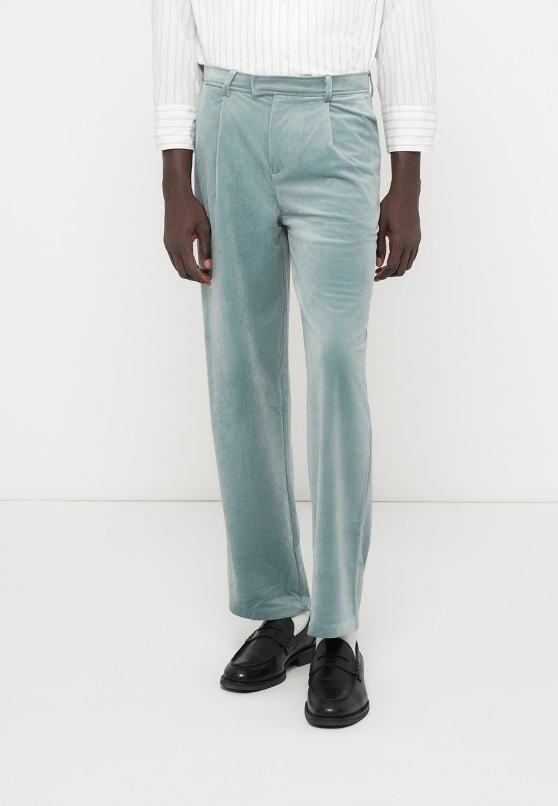 Calças de veludo numa tonalidade azul clara com uma textura suave, apresentando um ajuste ajustado, duas pregas à frente e bolsos laterais.