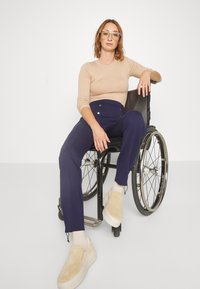 Beige ribbed long-sleeve top, high-waisted navy trousers with buttons, beige slip-on shoes, seated in a black wheelchair with metal wheels.
