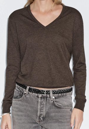 Femme portant un pull marron à manches longues et col en V rentré dans un jean gris avec une ceinture noire tressée et un bracelet en argent au poignet gauche.