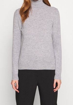 Kvinde iført en lysegrå langærmet højhalset sweater og sorte bukser, der står foran en enkel lys baggrund.