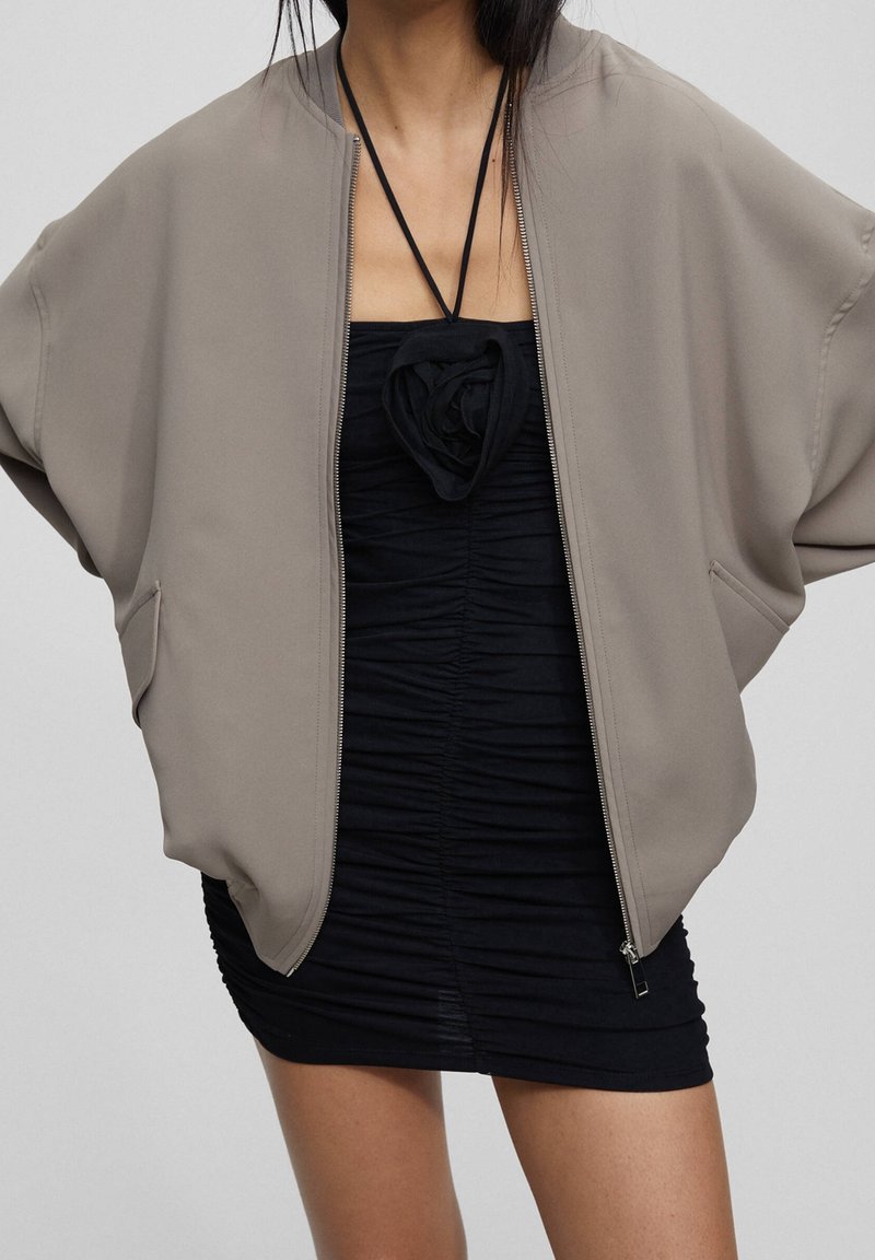 Veste bomber oversize beige avec deux poches latérales et une fermeture éclair, portée sur une mini-robe noire froncée avec un détail de décolleté floral.