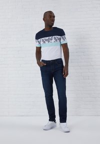 T-shirt à manches courtes avec un haut bleu marine, un corps blanc orné d'un motif de feuilles de palmier vertes et noires, associé à un jean bleu foncé et des baskets blanches.