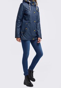 Tmavě modrá nepromokavá bunda s kapucí, zlatými knoflíky a stahovacími šňůrami. Nositelná přes modré džíny a černé šněrovací boty.