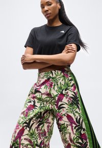 Mujer con cabello largo que lleva una camiseta negra y pantalones de pierna ancha con un estampado de pájaros tropicales y follaje, con rayas laterales verdes.