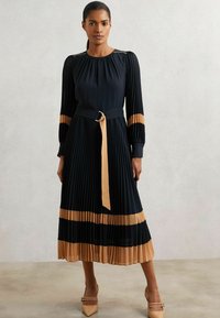 Kvinde iført en sort plisseret midi-kjole med lysebrune vandrette striber, et matchende bælte og lysebrune spidse hæle, der står foran en ensfarvet baggrund.