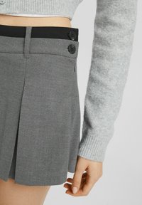 Gonna grigia plissettata con una vita nera, caratterizzata da due bottoni neri e una zip laterale, realizzata in un tessuto liscio con un design sartoriale.