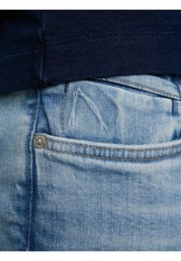 Vaqueros azul claro con una textura ligeramente desgastada, un bolsillo trasero visible y un botón metálico. La tela muestra un sutil desgaste y detalles de costura.